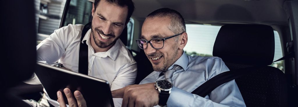 Smiling business colleagues using touchpad on the backs eat of the car. Focus is on businessman with glasses.